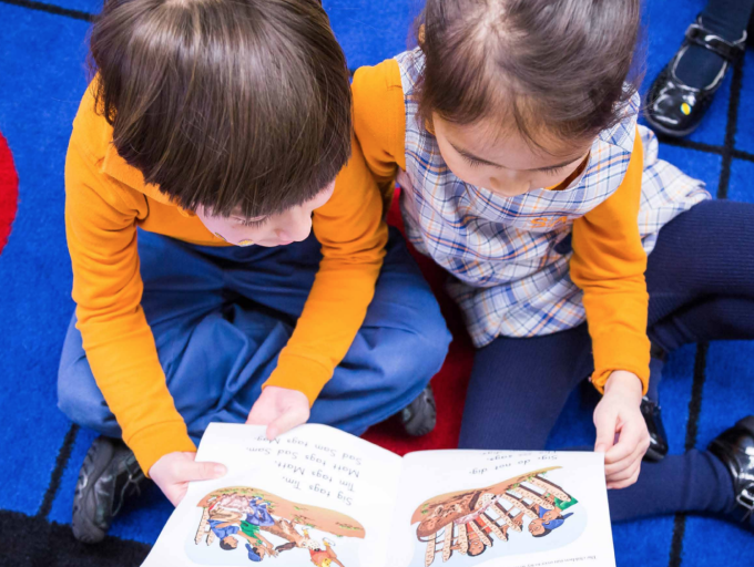 Children reading book together on colorful carpeted floor.