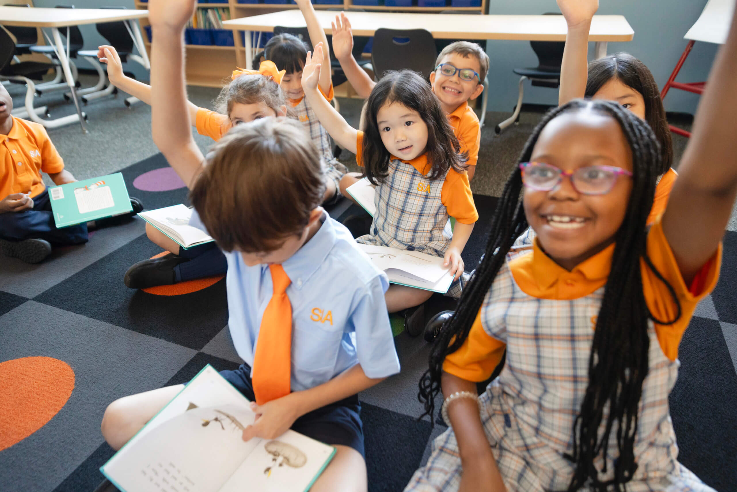 Children sitting cross-legged, raising hands, in classroom setting.