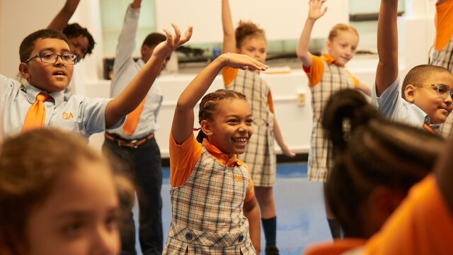 Children raise hands while smiling in a classroom setting.