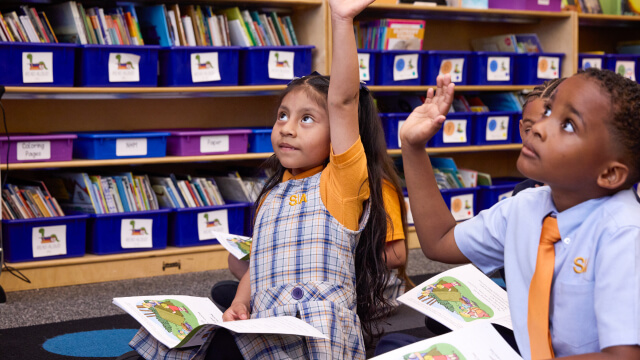 Children raise hands, sitting on floor, holding booklets. Classroom environment.