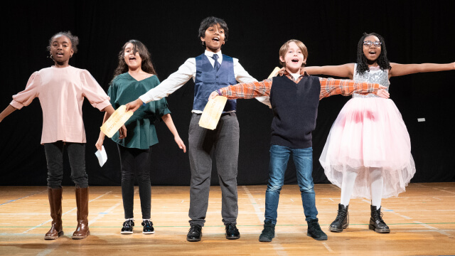 Children singing on stage, holding papers, in a theater.