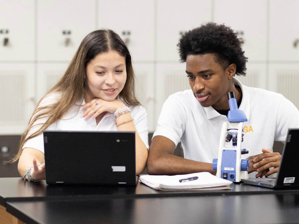 Students collaborate using laptops and microscope in classroom.
