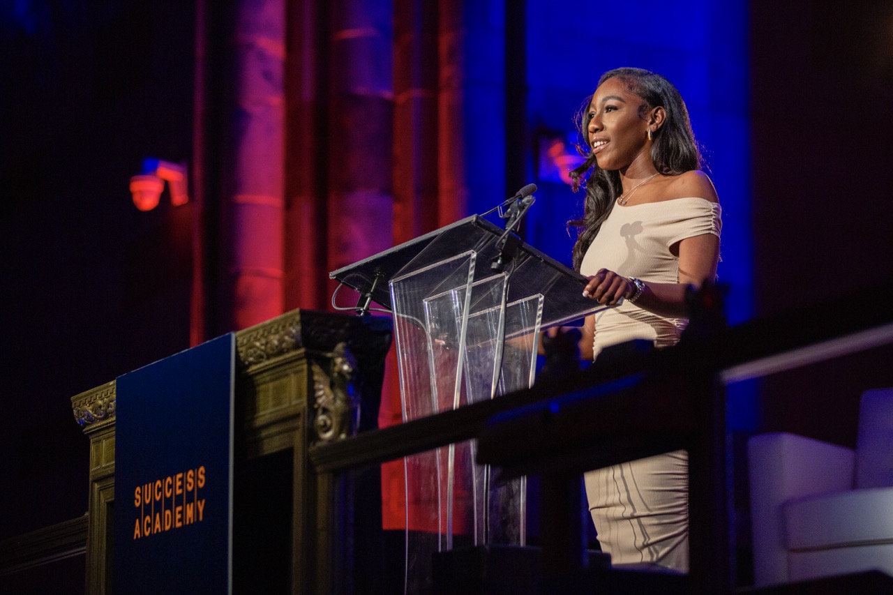 Woman speaking at podium during event, backdrop with colorful lights. Text on podium: 