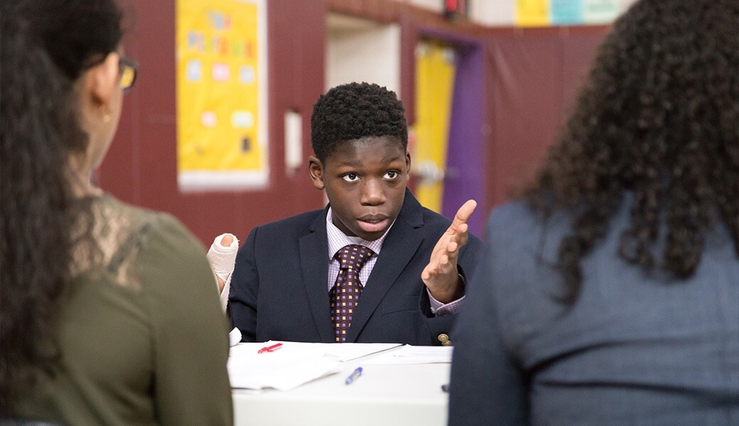 A young student in a suit passionately speaking during a discussion, gesturing with his hand while two individuals listen attentively.