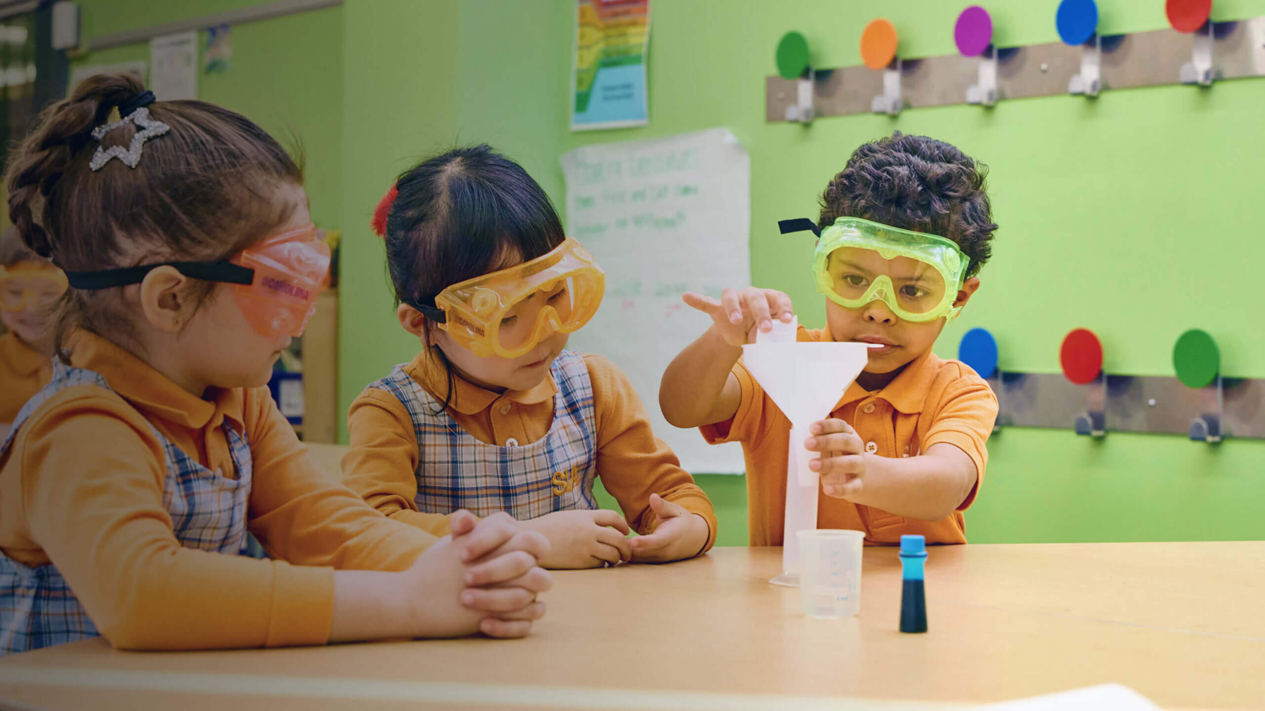 Kindergarten students wearing safety goggles while conducting a science experiment with funnels in a bright classroom.