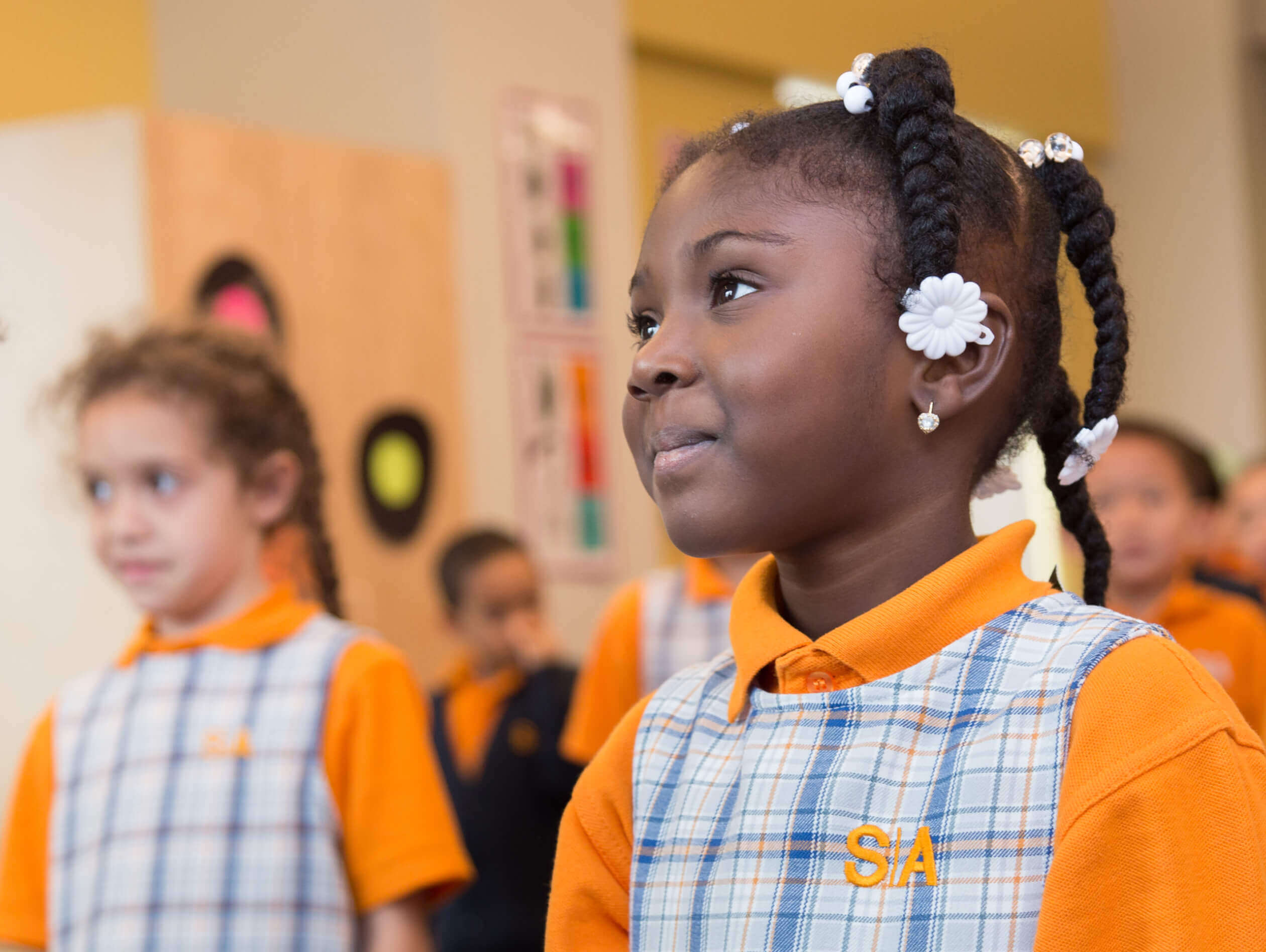 Smiling girl with braided hair and white flower accessories, wearing a school uniform in a classroom.