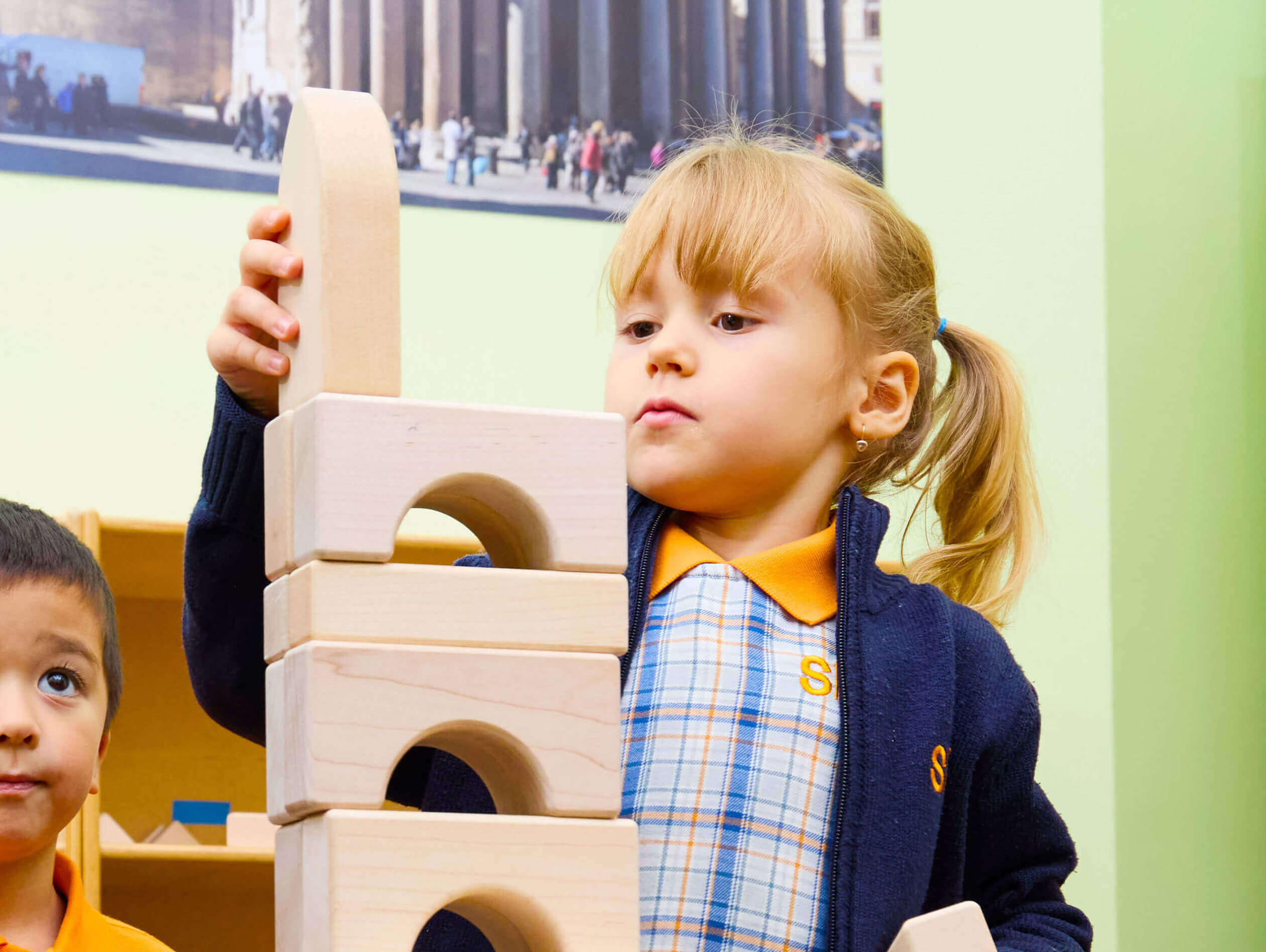Young girl carefully stacking wooden arch blocks in a classroom setting.