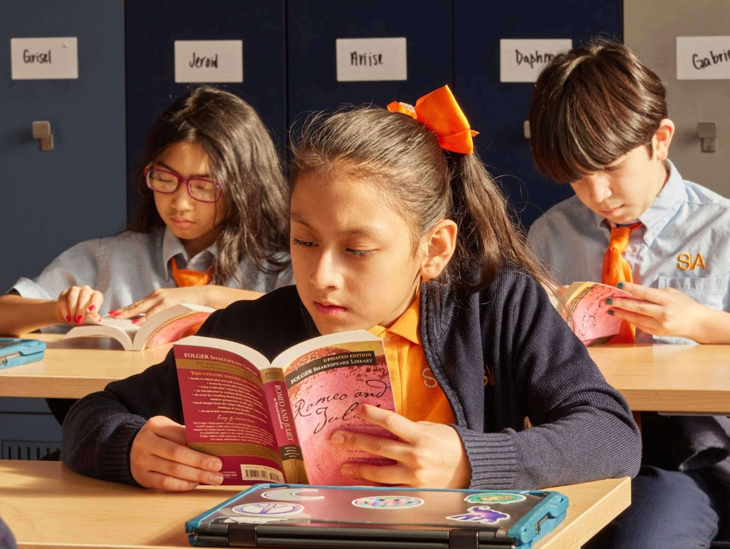 Three students reading books at a desk in a classroom.